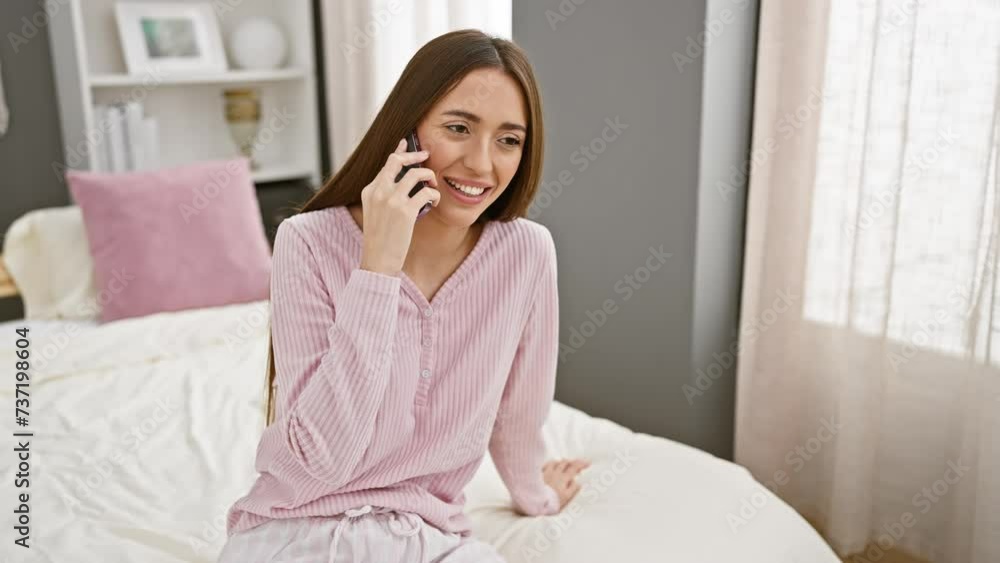 Young woman talking on smartphone while sitting in a cozy, stylish bedroom with white bedding and decorative pillows. Young woman talking on smartphone while sitting in a cozy, stylish bedroom with white bedding and decorative pillows.