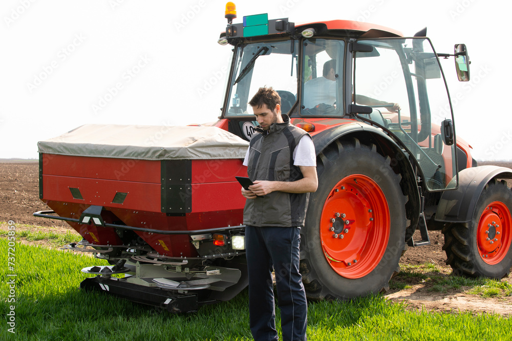 Obraz premium Young farm worker next to tractor using digital tablet. Tractor spreading artificial fertilizers in wheat field.
