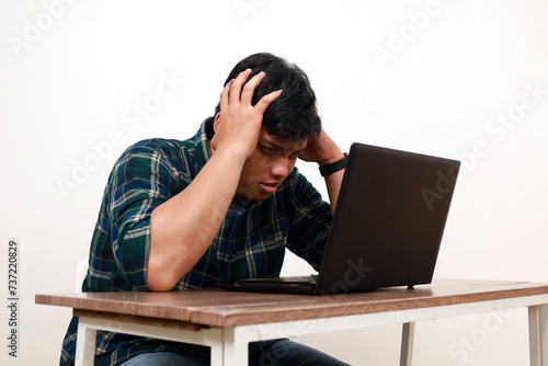 Stressed young asian college student sitting while holding his head with laptop on the desk