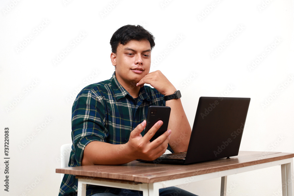 Happy young asian college student sitting while holding a cell phone and using laptop on the desk Happy young asian college student sitting while holding a cell phone and using laptop on the desk