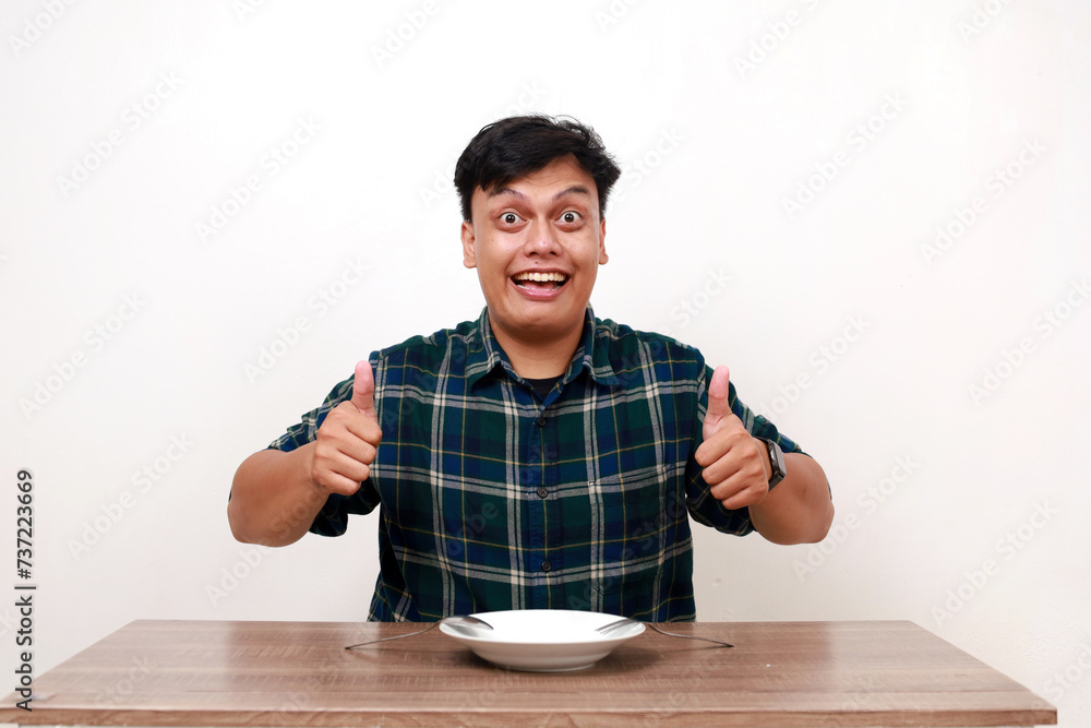 Excited young asian man showing thumbs up with empty plate on the table Excited young asian man showing thumbs up with empty plate on the table