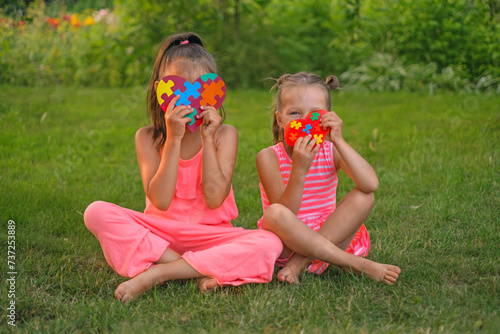 Happy playful girls with hearts made of puzzle pieces are sitting on the grass in the garden