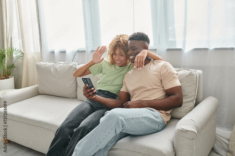 happy african american couple video chatting on a smartphone while sitting on couch together happy african american couple video chatting on a smartphone while sitting on couch together