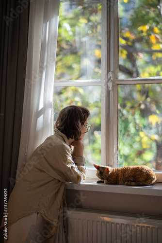 Pensive lonely woman in melancholy looking at window, thinking of life, spending weekend, pastime at home in summer sunny day. Cat Devon Rex lying on windowsill, warming and sleeping. Loneliness. 