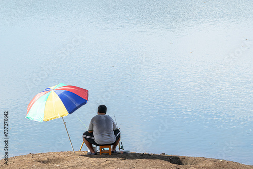 A man is sitting fishing on the bank of a river. man sitting under an umbrella. Man, relaxing and fishing by the lakeside.