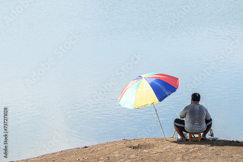 A man is sitting fishing on the bank of a river. man sitting under an umbrella. Man  relaxing and fishing by the lakeside.