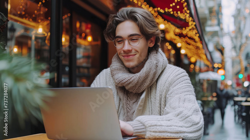 Cheerful young man with glasses working on laptop in festive cafe, urban winter charm. Generative AI