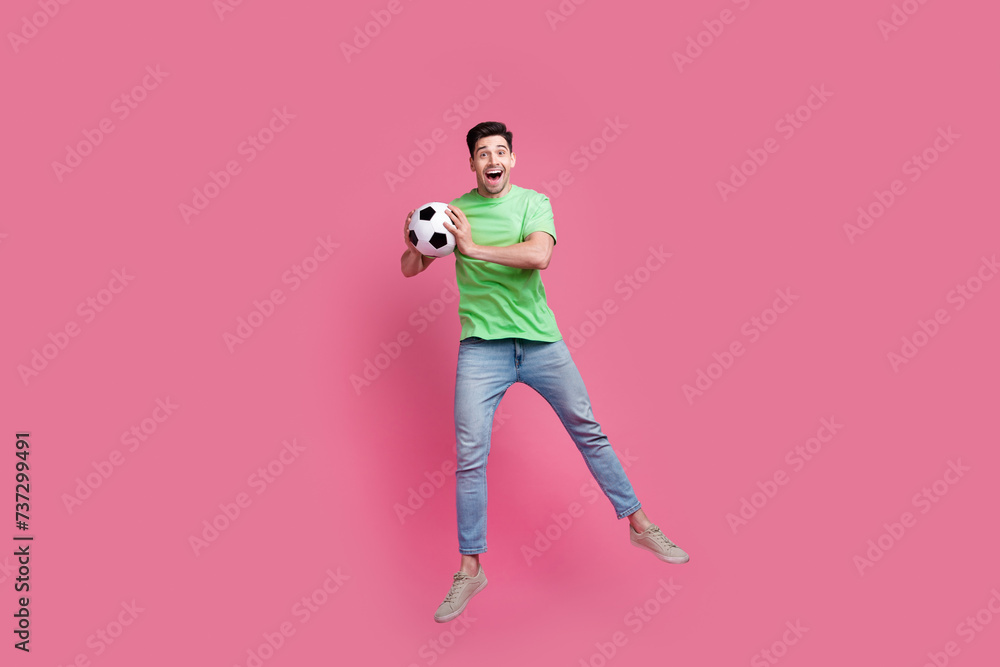 Full length body photo of young sportsman jumping wearing green t shirt catch ball playing soccer game isolated on pink color background Full length body photo of young sportsman jumping wearing green t shirt catch ball playing soccer game isolated on pink color background