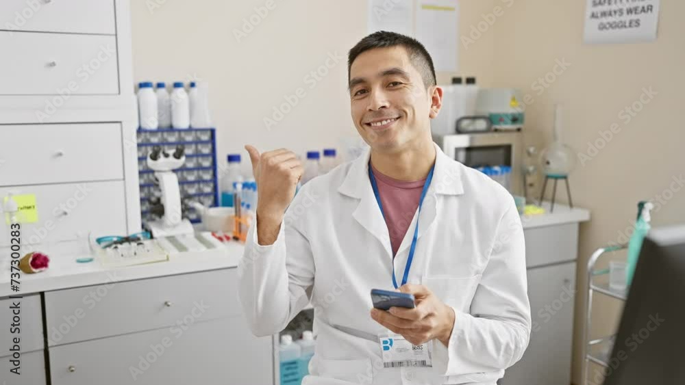 Cheerful young hispanic male scientist in a lab uniform, confidently pointing his thumb up, holding a smartphone, sporting an open-mouth smile in the laboratory Cheerful young hispanic male scientist in a lab uniform, confidently pointing his thumb up, holding a smartphone, sporting an open-mouth smile in the laboratory