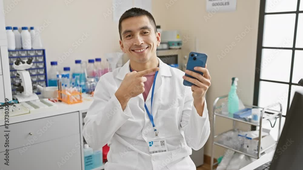 Joyful young hispanic man scientist in uniform excitedly pointing with hand and finger, showing an idea on his smartphone at the lab, exuding a confident, optimistic expression. Joyful young hispanic man scientist in uniform excitedly pointing with hand and finger, showing an idea on his smartphone at the lab, exuding a confident, optimistic expression.