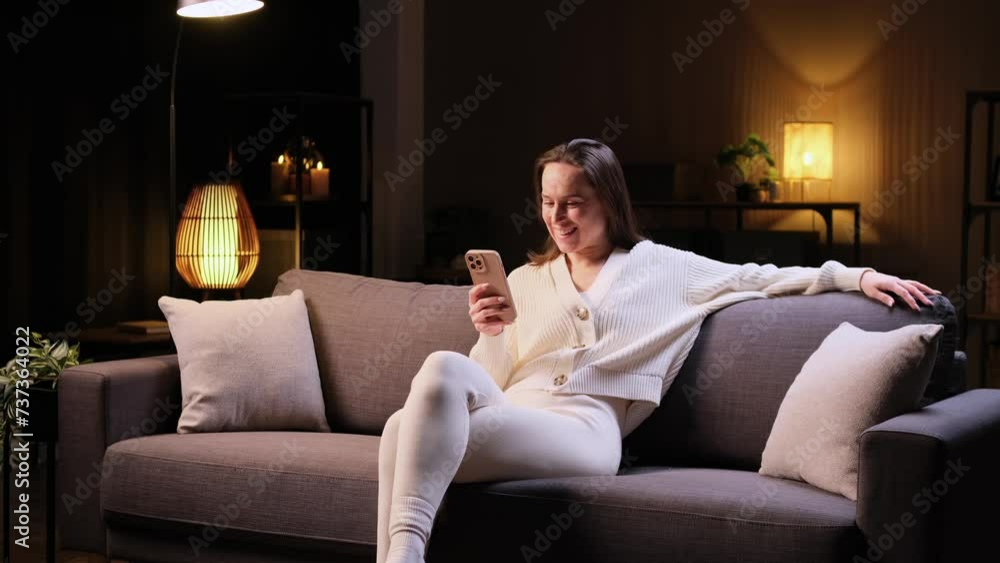 Smiling Caucasian woman surfing the internet using phone, relaxing at evening home, sitting on sofa. Enjoying leisure activity, online entertainment. Smiling Caucasian woman surfing the internet using phone, relaxing at evening home, sitting on sofa. Enjoying leisure activity, online entertainment.