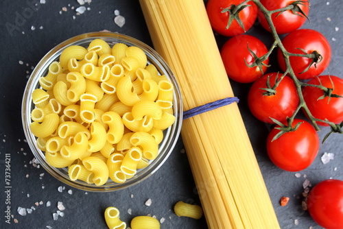 On a black background lies a long spaghetti, a glass bowl with small pasta and a sprig of cherry tomatoes sprinkled with large crystals of sea salt.