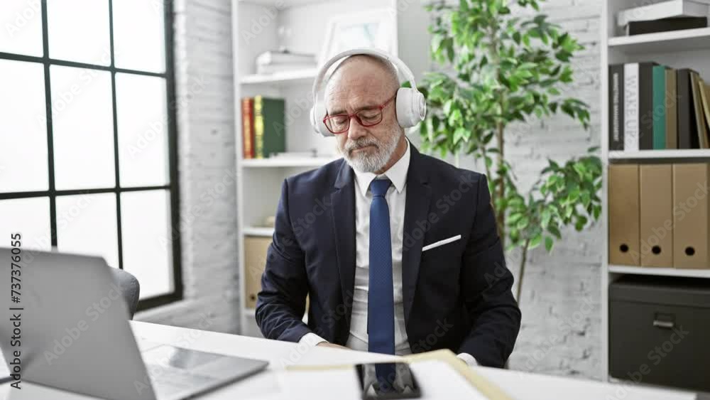 A mature man in a suit engages with technology in a modern office, wearing headphones and using a smartphone. A mature man in a suit engages with technology in a modern office, wearing headphones and using a smartphone.