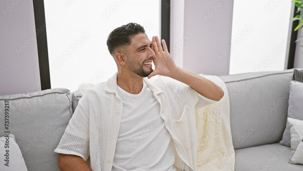 Agitated young arab man, seated on the home sofa, yells and screams wildly off to the side, hand cupping mouth in fiery display of vocal expression Agitated young arab man, seated on the home sofa, yells and screams wildly off to the side, hand cupping mouth in fiery display of vocal expression