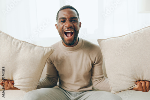 Relaxed African American man sitting on a comfortable sofa in his modern living room, smiling and holding a pillow He is enjoying a peaceful weekend at home, thinking and watching something online