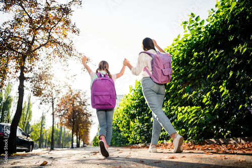 Full size rear portrait of two funky carefree sisters hold arms carry rucksack walk park sunlight outdoors