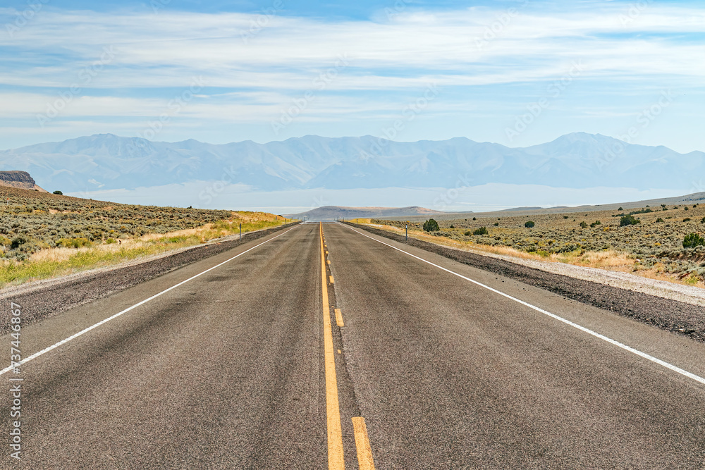 Deserted asphalt highway through the Nevada desert summer landscape with haze covered mountain range and blue sky with clouds. USA.