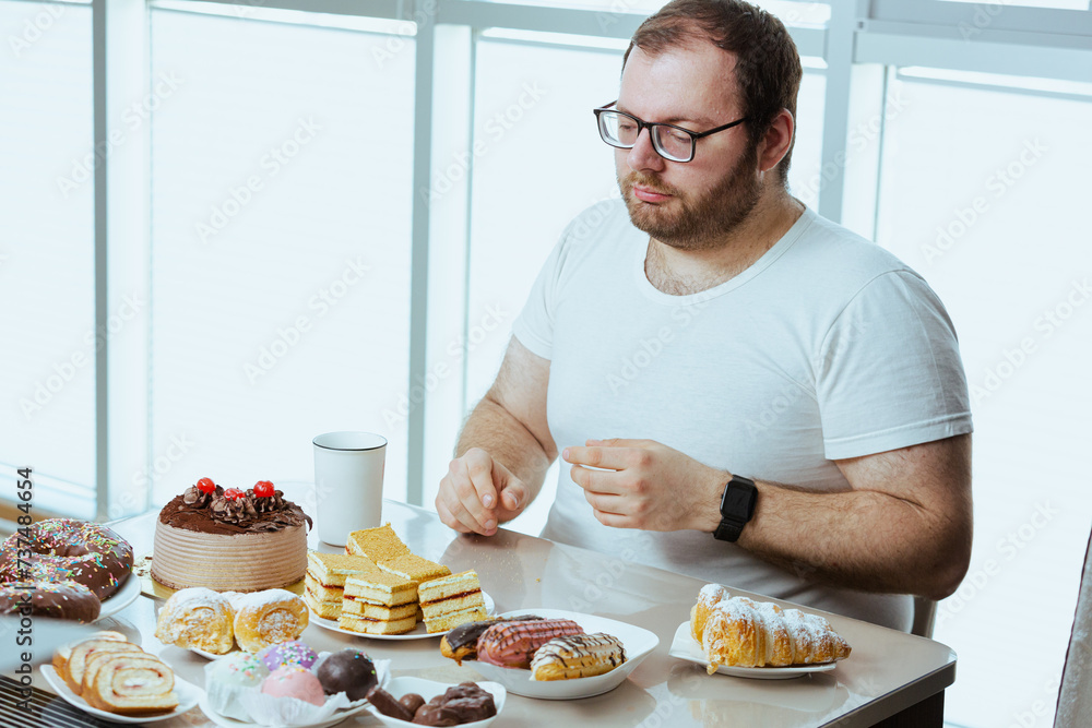Man sitting in front of a table full of sweet food, eating pastry with cream. Fat man sugar addict, can't say no and stop eating, weight loss problem. Depressed man eats a lot of unhealthy meal. Man sitting in front of a table full of sweet food, eating pastry with cream. Fat man sugar addict, can't say no and stop eating, weight loss problem. Depressed man eats a lot of unhealthy meal.