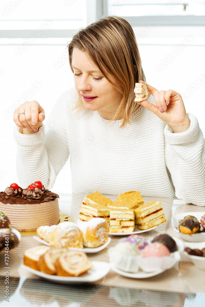 Woman eating pastry with cream, picking what to eat sitting beside table full of sweets. Diet is over, sugar addiction and problem with diabetes. Woman eating pastry with cream, picking what to eat sitting beside table full of sweets. Diet is over, sugar addiction and problem with diabetes.