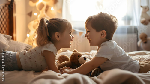 Two cute children laughing at each other while lying in their bed. Sibling love. Brother and sister holding a teddy bear and looking at each other.