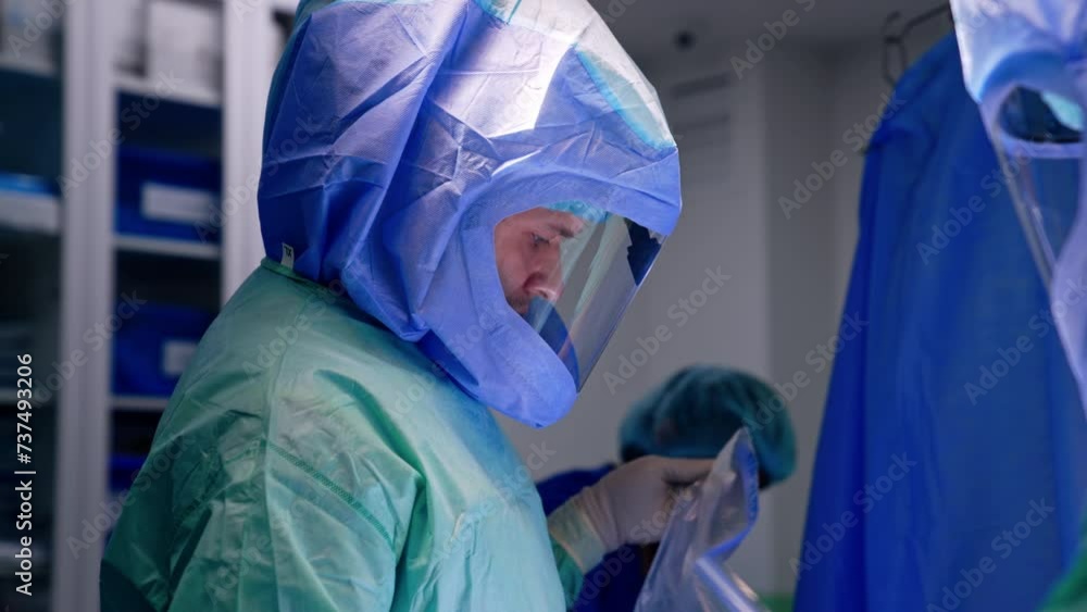 Male doctor in robe and helmet holding a piece of plastic. Side view. Medics working in modern surgery room. Male doctor in robe and helmet holding a piece of plastic. Side view. Medics working in modern surgery room.