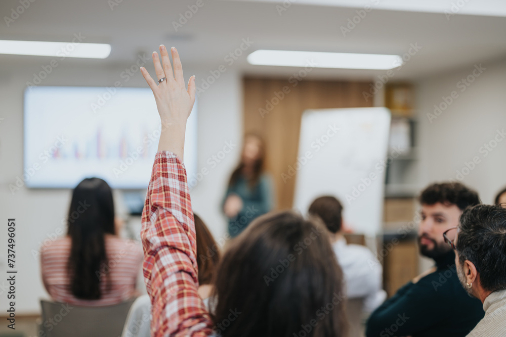 © qunica.com - An attentive participant raises their hand to ask a question during a business seminar, with fellow attendees and presenter in the background.