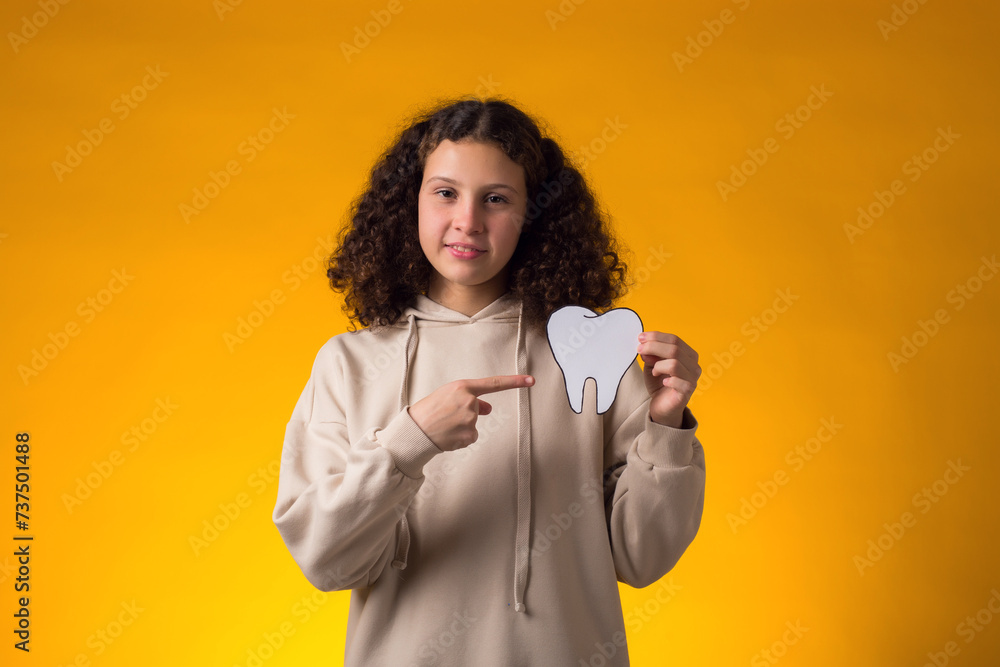 Girl holding papercraft tooth and pointing finger at it over yellow background. Dental health concept Girl holding papercraft tooth and pointing finger at it over yellow background. Dental health concept