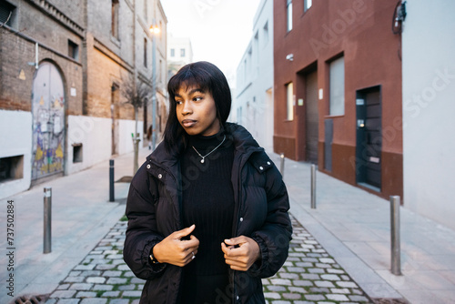 Portrait of a cheeful young black woman with a coat walking on a narrow street. © Jordi Salas