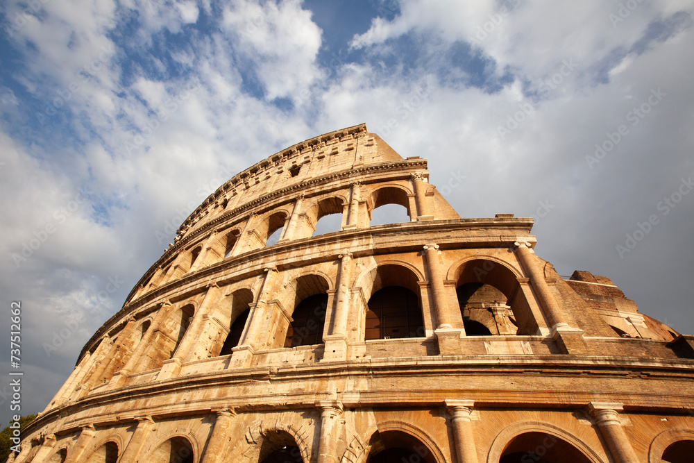 Colosseum in Rome, framed from a low angle, showcasing the upper section against a backdrop of partly cloudy skies