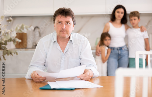 Wistful father looking through papers sitting in the kitchen, his wife and children looking at him with compassion standing behind