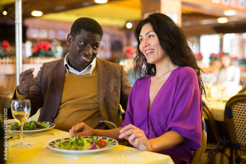 Cheerful african american man flirting with attractive asian woman during dinner in restaurant..