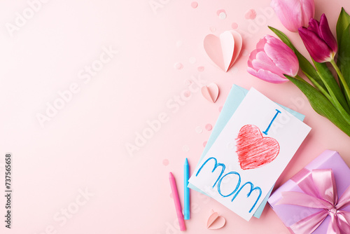 Tender tribute: crafted affection for mother’s day. Top view of pink tulips, child's drawing expressing love for mom, pink crayon, gift box on light pink background with space for greetings