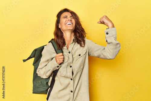 Middle aged woman ready for travel with backpack on yellow raising fist after a victory, winner concept.