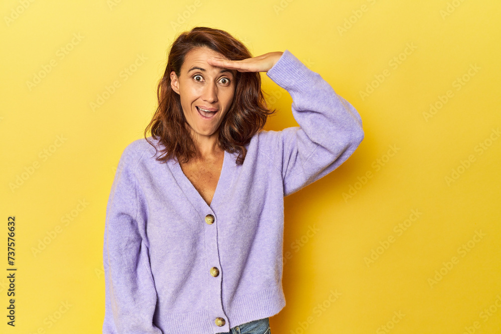 Fototapeta premium Middle-aged woman on a yellow backdrop shouts loud, keeps eyes opened and hands tense.