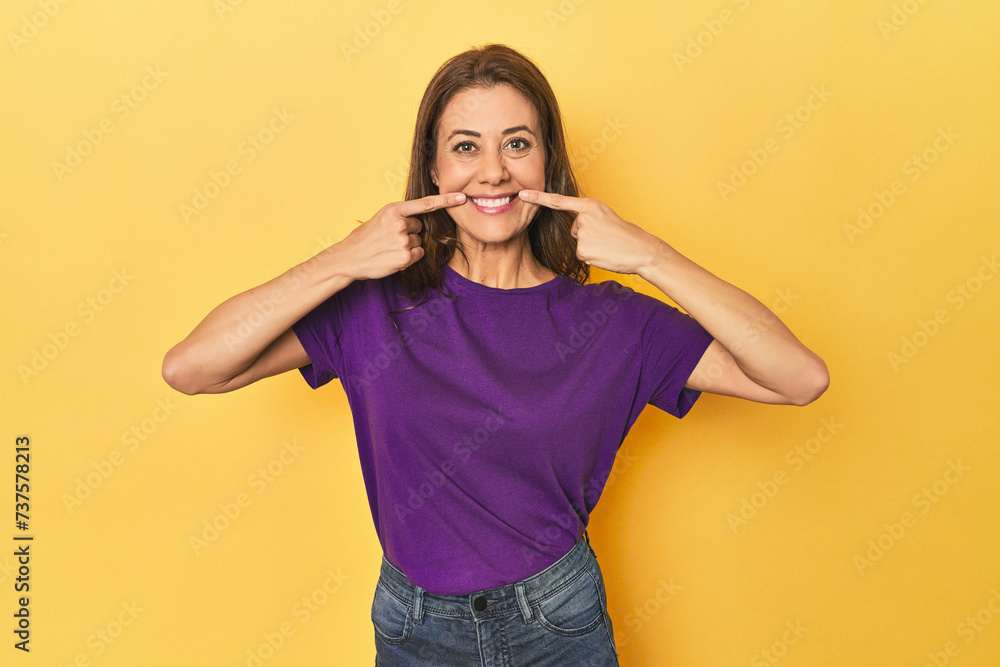 Middle-aged caucasian woman on yellow smiles, pointing fingers at mouth. Middle-aged caucasian woman on yellow smiles, pointing fingers at mouth.