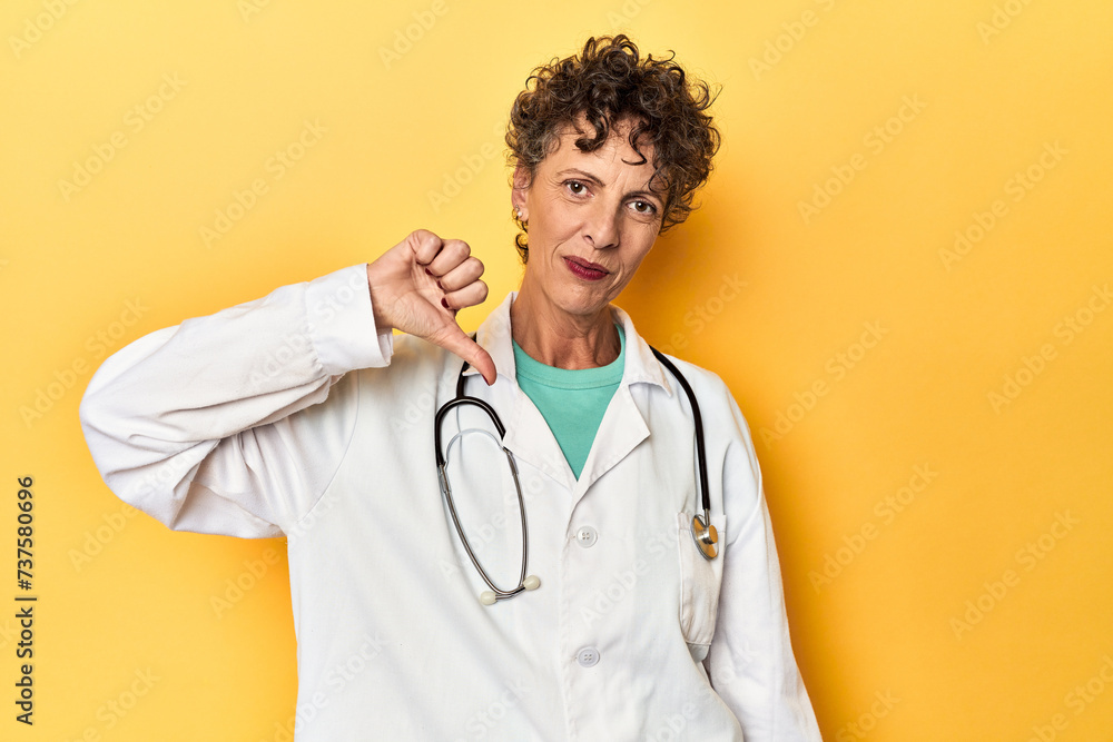 Caucasian mid-age female doctor on yellow studio showing a dislike gesture, thumbs down. Disagreement concept. Caucasian mid-age female doctor on yellow studio showing a dislike gesture, thumbs down. Disagreement concept.