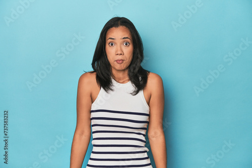 Filipina young woman on blue studio shrugs shoulders and open eyes confused.