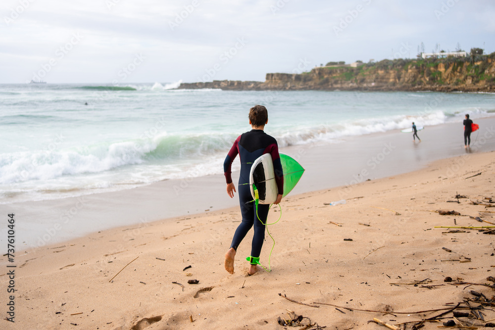 Back view of anonymous surfer in wetsuit with surfboard walking along sea waves. Active surfer in swimwear walking on sandy beach. Embodying the concept of a summer vacation. Back view of anonymous surfer in wetsuit with surfboard walking along sea waves. Active surfer in swimwear walking on sandy beach. Embodying the concept of a summer vacation.