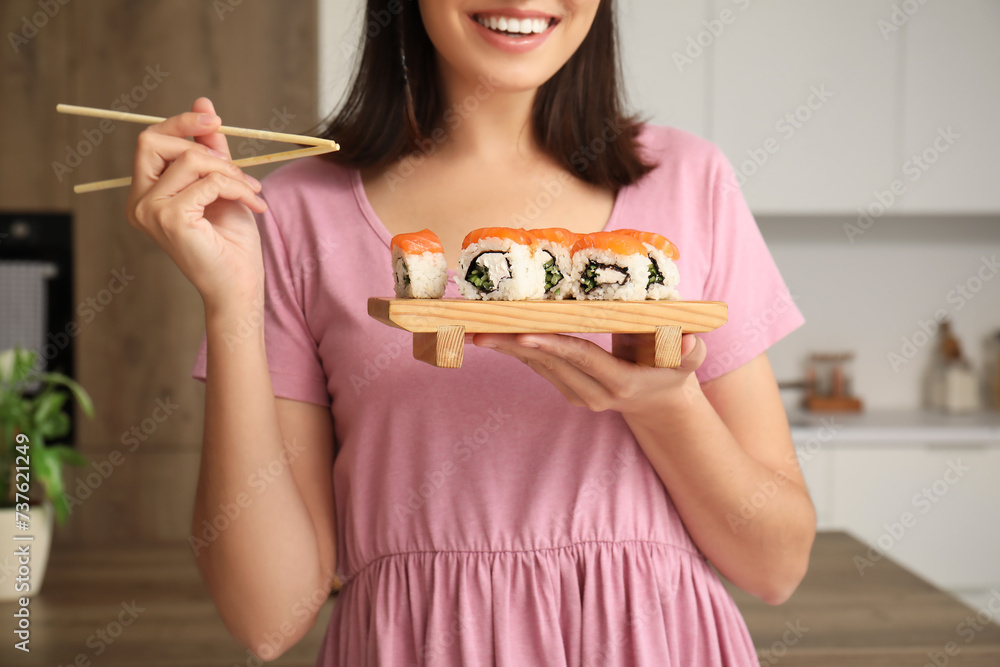 Fototapeta premium Young woman eating tasty sushi rolls in kitchen
