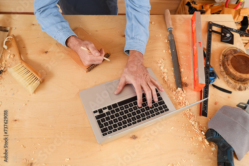 Mature carpenter working with laptop at table in shop, top view