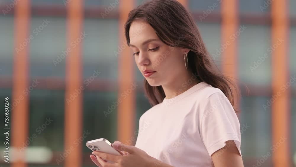 A young woman is focused on her smartphone, possibly waiting for a message or a friend, with a blurred urban backdrop setting the scene. A young woman is focused on her smartphone, possibly waiting for a message or a friend, with a blurred urban backdrop setting the scene.