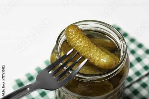 Fork with pickled cucumber over jar on white table, closeup
