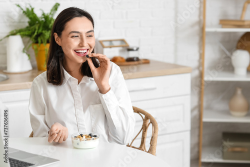 Happy young woman with blueberries and tasty yoghurt at table in kitchen