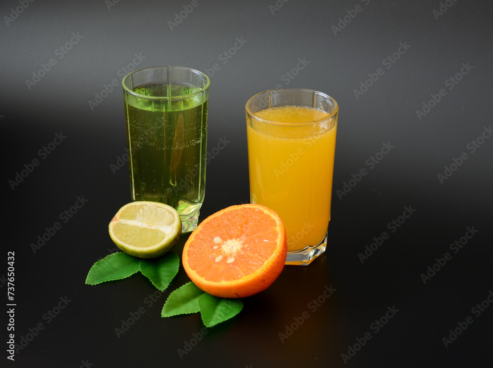 Two tall glasses with different citrus juices on a black background, next to the halves of ripe orange and lime with leaves. Two tall glasses with different citrus juices on a black background, next to the halves of ripe orange and lime with leaves.