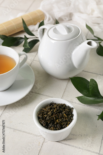 Teapot and bowl with dry leaves on white tile background, closeup