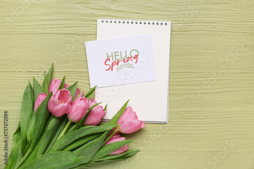 Greeting card with text HELLO SPRING, notebook and beautiful pink tulips on green wooden background