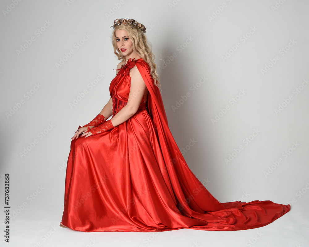 Full length portrait of beautiful blonde model dressed as ancient mythological fantasy goddess in flowing red silk toga gown, crown. Seated pose sitting on chair. isolated on white studio background.