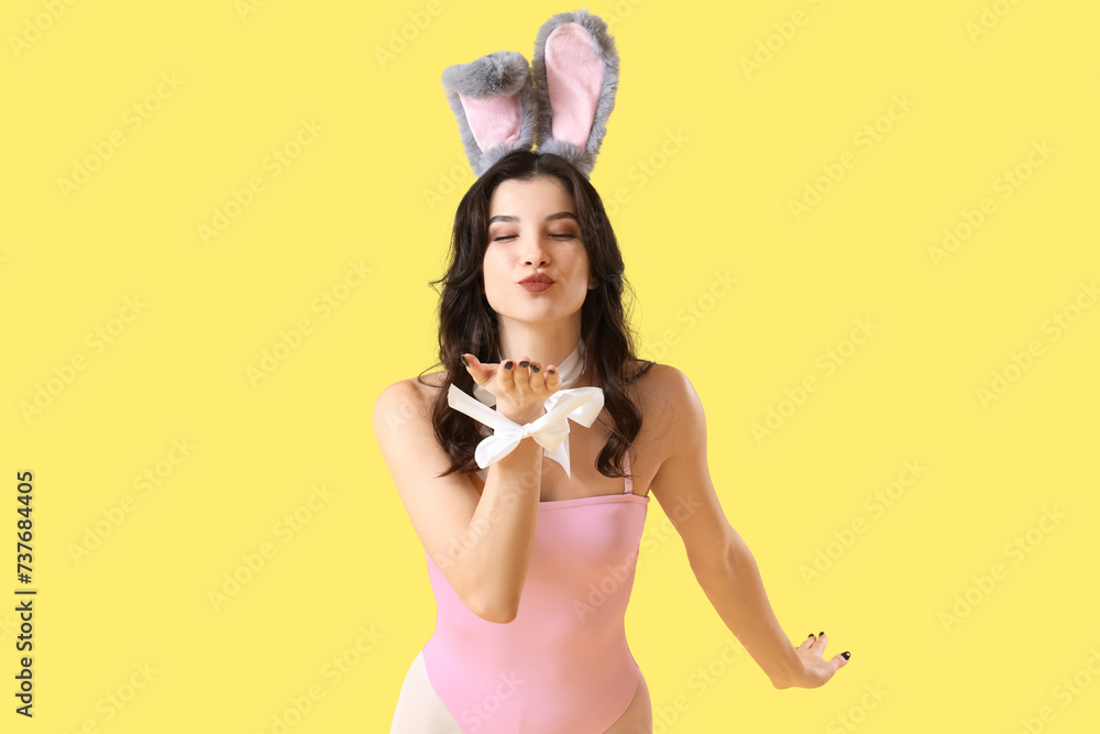 Sexy young woman in bunny costume blowing kiss on yellow background. Easter celebration