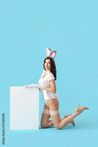 Sexy young woman in bunny costume with blank poster on blue background. Easter celebration