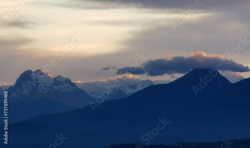 Le cime delle montagne innevate al tramonto nel cielo plumbeo invernale e nuvole bordate di rosso dalla luce del sole © GjGj